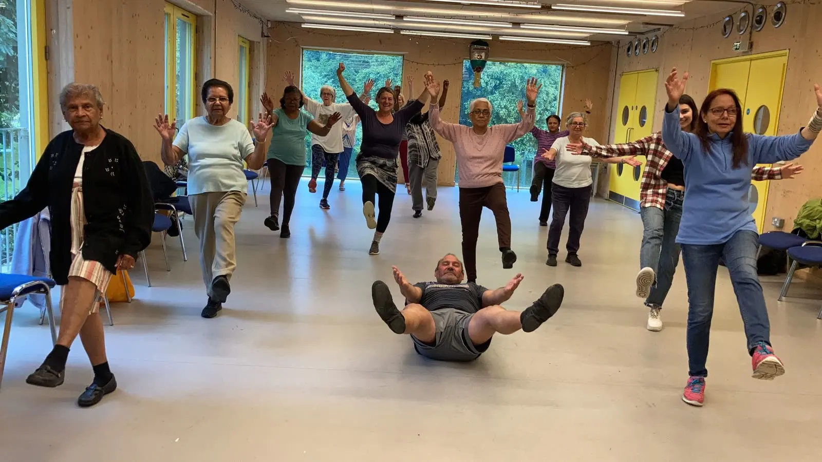 A class of older adults balancing on one leg with arms raised in a bright hall